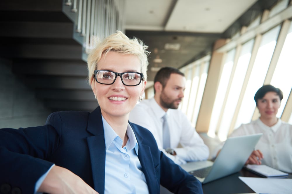 blonde with short hairstyle and glasses, business woman on meeting, people group in background at modern bright office indoors