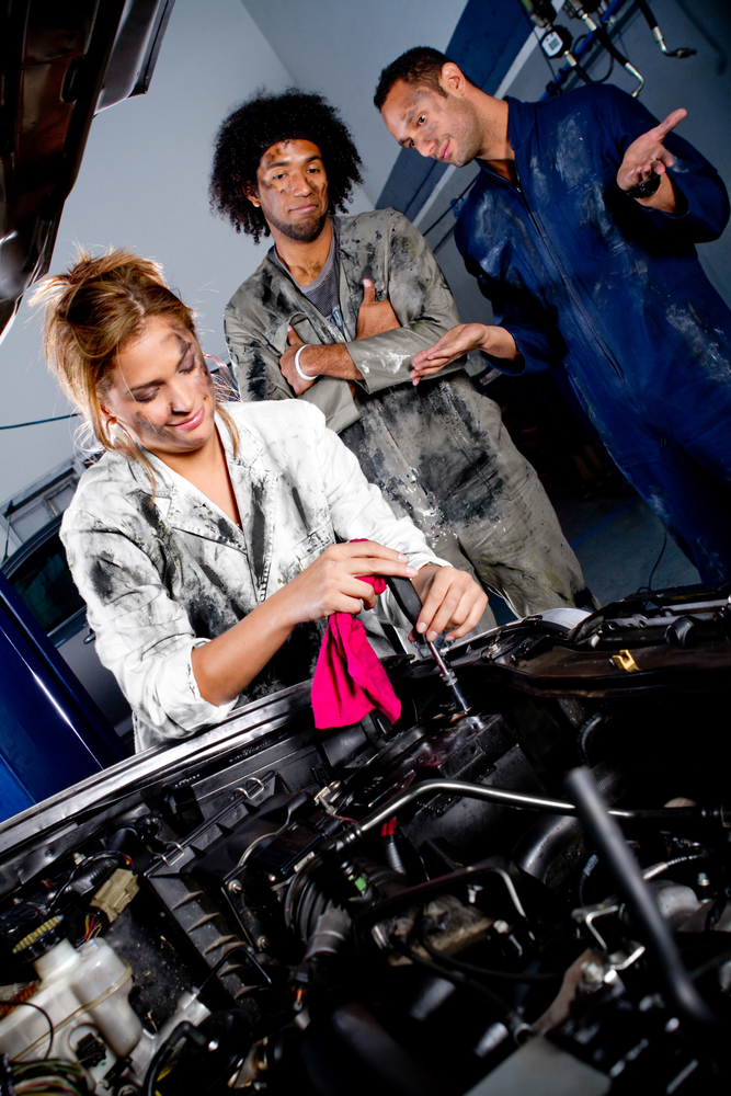 Woman working on a car while men look on.
